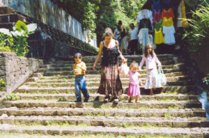 A woman and young children leave a shadow-filled walkway, descending steps into bright sunlight.