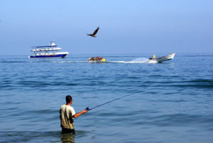 A vacationer wades into the surf to fish while a boat pulls a long yellow 'banana' — an inflatable rubber tube — with laughing riders. In Mexico's Jaltemba Bay, Easter holidays are akin to a family-friendly Mardi Gras. © Christina Stobbs, 2011