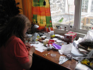 As Martha works on her feather art, she can look out a window at the birds screeching in the late morning light. El loro Lorenzo keeps close watch over the situation.