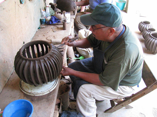 José María Alejos Madrigal cuts a slice from a cactus-shaped pot. In the background, his wife works on a ceramic pig.
