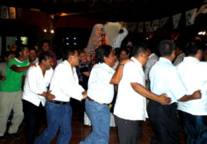 The bride and groom stand on chairs while Alvin holds Arlene's bridal "train" (formed by tucking a rebozo into the waist of her skirt) aloft to form a bridge. First single women form a conga line that passes beneath the bridge while music plays. They then try to catch the bride's bouquet. Here, Alvin has removed his wife's satin garter and single men dance in line while waiting for him to throw it, as is done in most Mexican wedding parties. © Alvin Starkman 2008