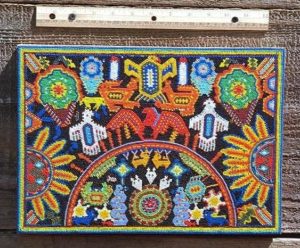 This beadwork votive painting depicts elements of Huichol symbolism and cosmology such as the white candles, two orange jaguars, green and blue peyote buds, and the eagle. Inside the circle, a scorpion serpent and various portrait of the sacred deer can be seen. © Robert Otay, 1997