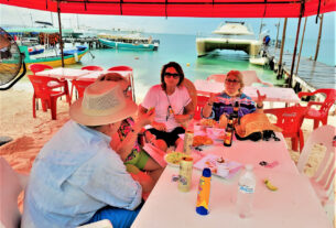 People at table by boats: Island visitors indulging in grilled fresh fish, house made tortillas, and, of course, lobster at the Miramar Lobster House. You can even get your photo taken there and pasted on a bottle to tequila to take home as a souvenir.© 2021 Jane Simon Ammeson