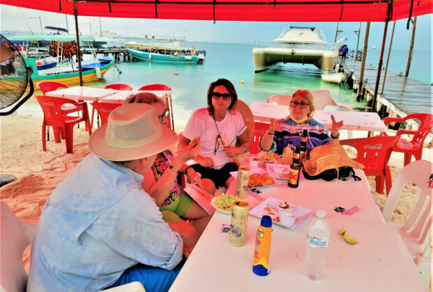 People at table by boats: Island visitors indulging in grilled fresh fish, house made tortillas, and, of course, lobster at the Miramar Lobster House. You can even get your photo taken there and pasted on a bottle to tequila to take home as a souvenir.© 2021 Jane Simon Ammeson